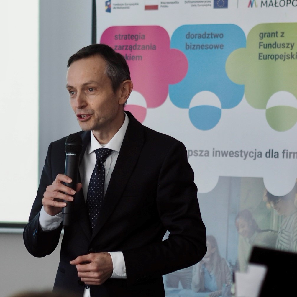 Elegancko ubrany mężczyzna w średnim wieku mówi do mikrofonu na tle baneru projektu "Pracownicy - najlepsza inwestycja dla firmy".