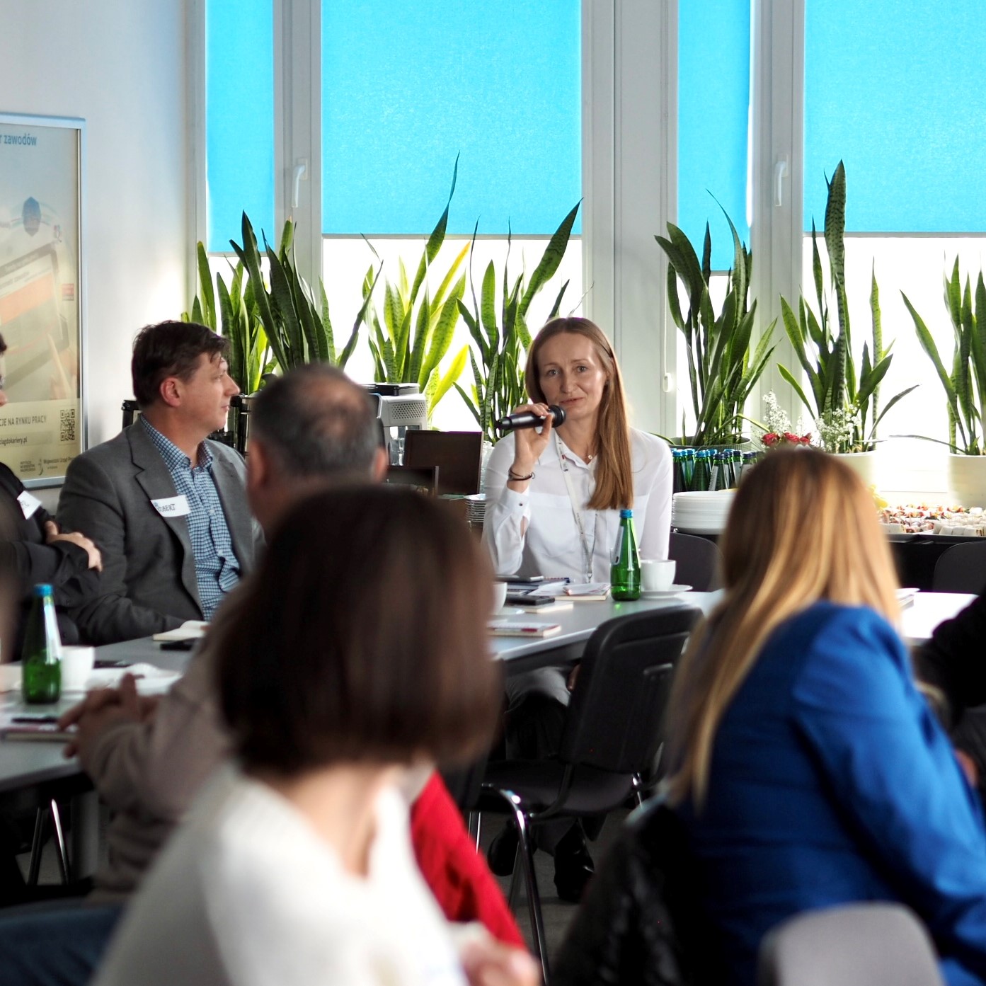 Młoda kobieta siedząca w grupie ludzi na spotkaniu mówi do mikrofonu. Wokół widoczne są twarze ludzi zwróconych w jej kierunku.