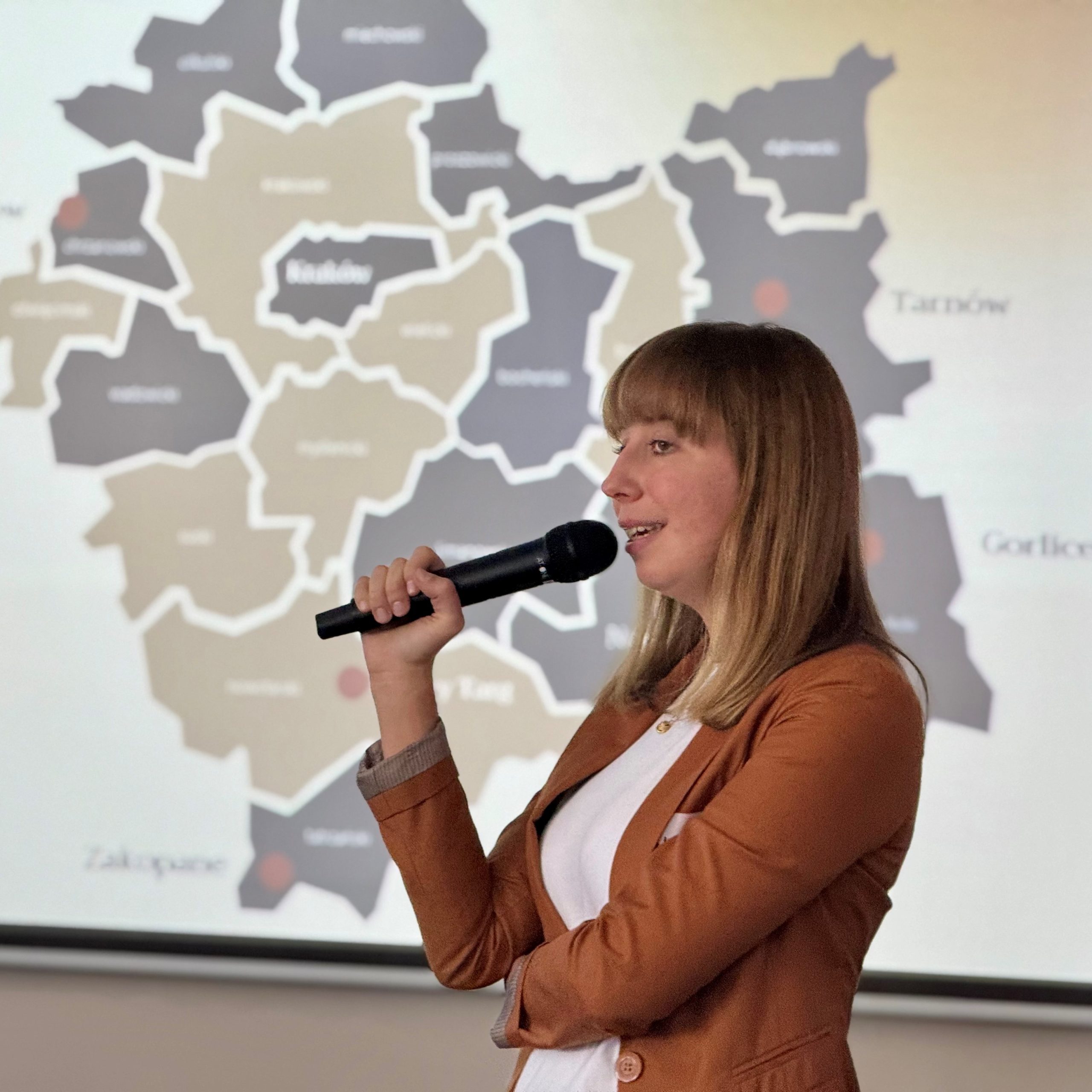 Młoda kobieta z jasnymi włosami mówi do mikrofonu na tle mapy województwa małopolskiego.