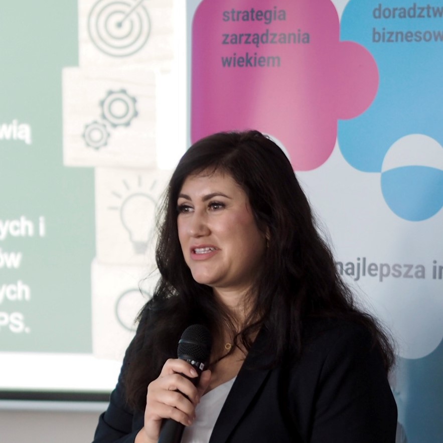 Młoda kobieta z długimi ciemnymi włosami mówi do mikrofonu na tle prezentacji.