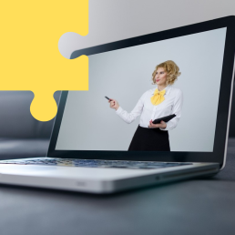 Na stole leży otwarty laptop. Na jego ekranie widać elegancko ubraną kobietę, która gestykuluje.