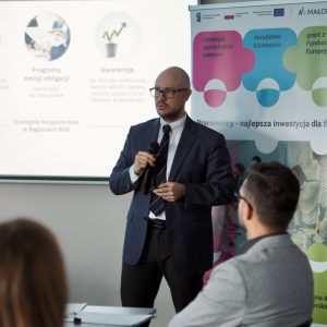 Elegancko ubrany mężczyzna mówi do grupy siedzących ludzi. W tle widać prezentację.