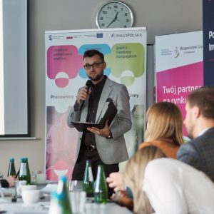 Młody mężczyzna w czarnej koszuli i szarej marynarce zwraca się do uczestników spotkania. Stoi na tle baneru projektu "Pracownicy - najlepsza inwestycja dla firmy".