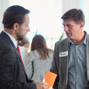 Dwaj elegancko ubrani mężczyźni rozmawiają ze sobą. Jeden z nich ma identyfikator z imieniem Robert.