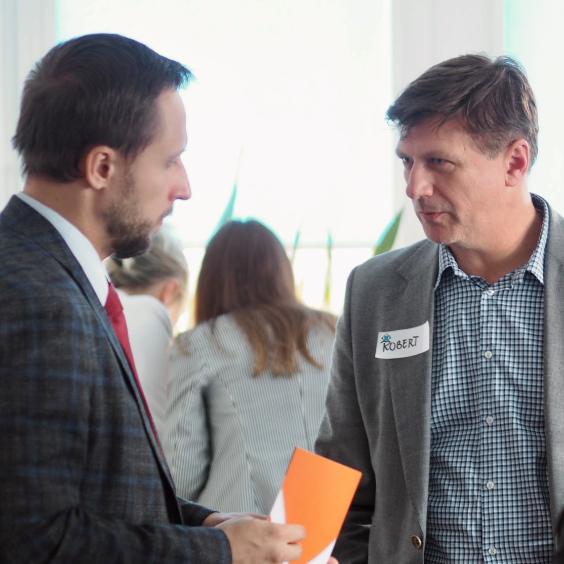 Dwaj elegancko ubrani mężczyźni rozmawiają ze sobą. Jeden z nich ma identyfikator z imieniem Robert.