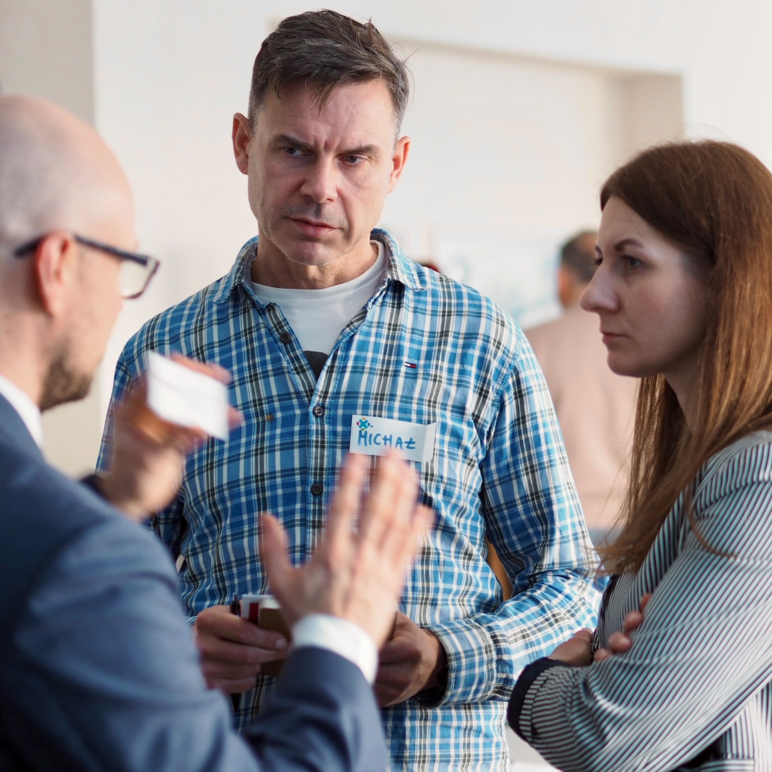 Dwóch mężczyzn i kobieta rozmawiają ze sobą, wymieniając się wizytówkami. Na piersi mężczyzny stojącego na wprost widoczny jest identyfikator z imieniem Michał.