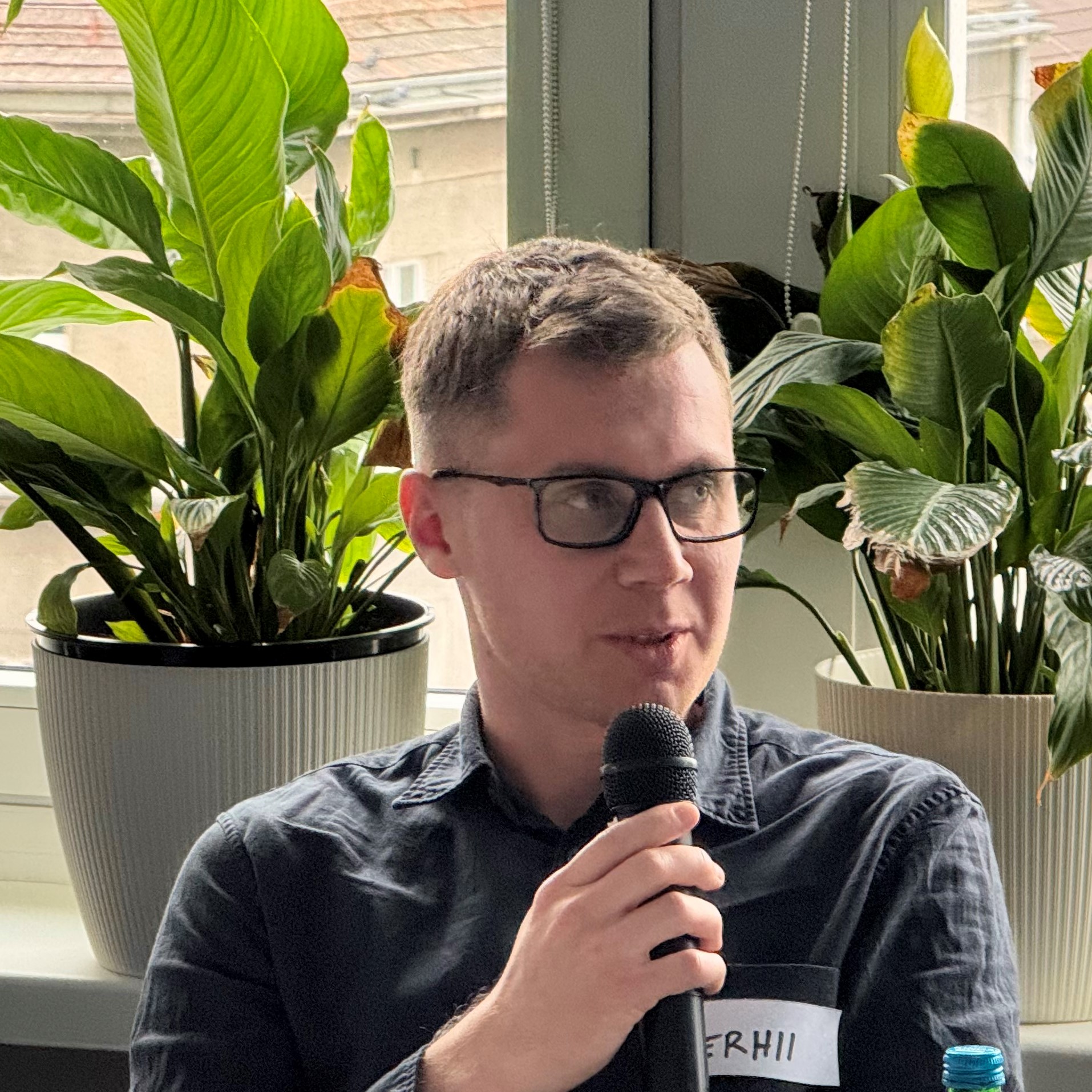 Młody mężczyzna w ciemnej koszuli mówi do mikrofonu, siedząc przy stole.