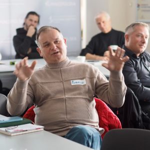 Mężczyzna w jasnym golfie gestykuluje, mówiąc do ludzi na sali. Na piersi ma identyfikator z imieniem Tomasz.