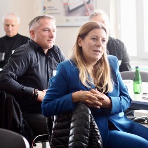 Blondynka z długimi włosami i w niebieskim garniturze mówi do uczestników spotkania. Za nią siedzi mężczyzna w czarnej koszuli.