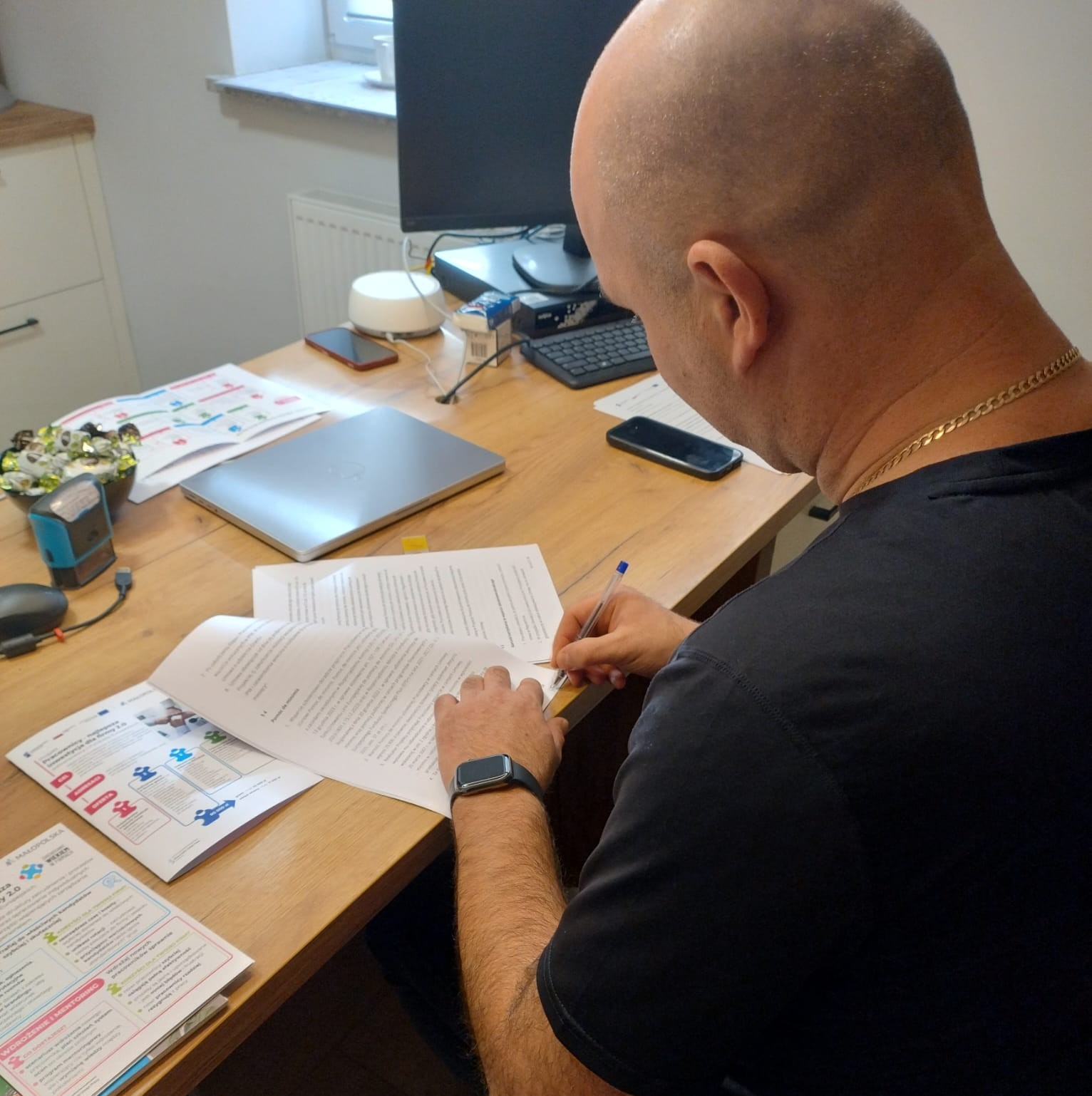 Łysy mężczyzna w ciemnej koszulce podpisuje dokumenty przy biurku.