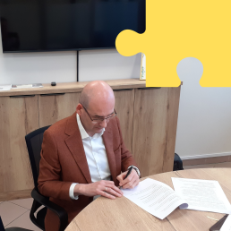 Łysy mężczyzna w okularach i brązowej marynarce podpisuje dokumenty.