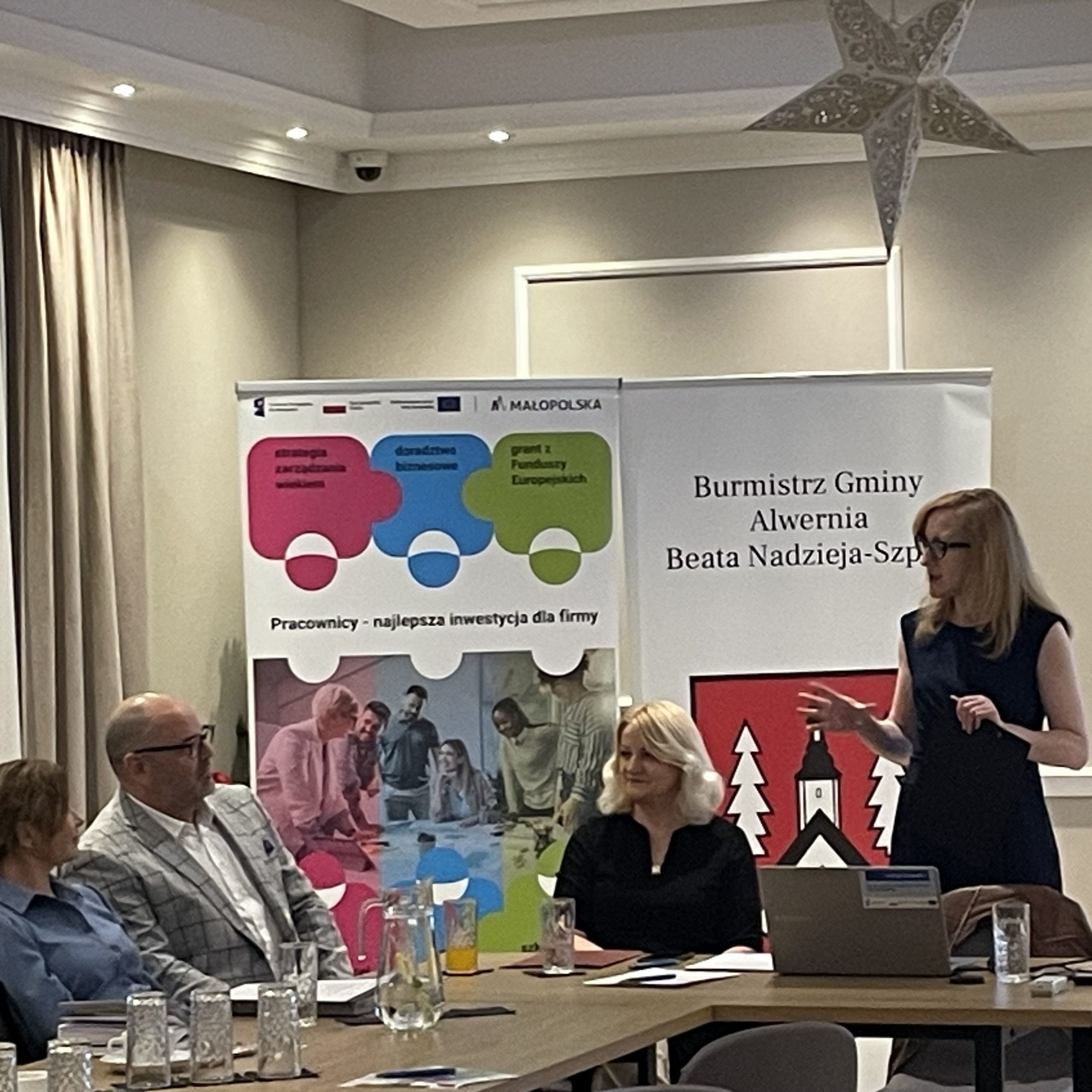 Stojąca kobieta w okularach rozmawia z parą, która siedzi przy stole.