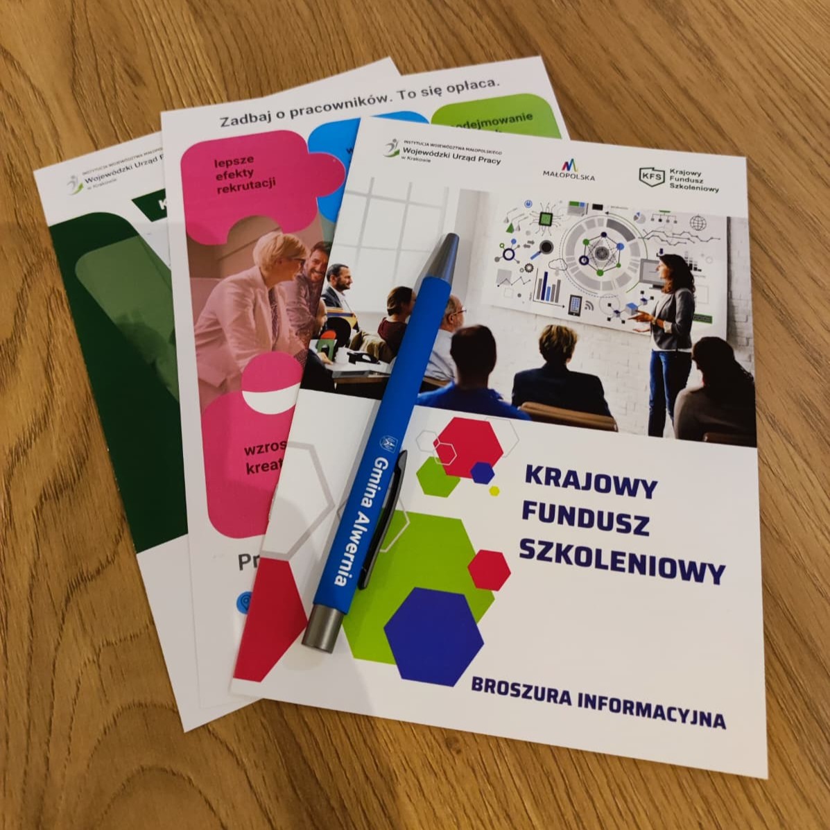 Zbliżenie na materiały informacyjne na temat projektu "Pracownicy - najlepsza inwestycja dla firmy" oraz Krajowego Funduszu Szkoleniowego. Na nich leży niebieski długopis z napisem: Gmina Alwernia.