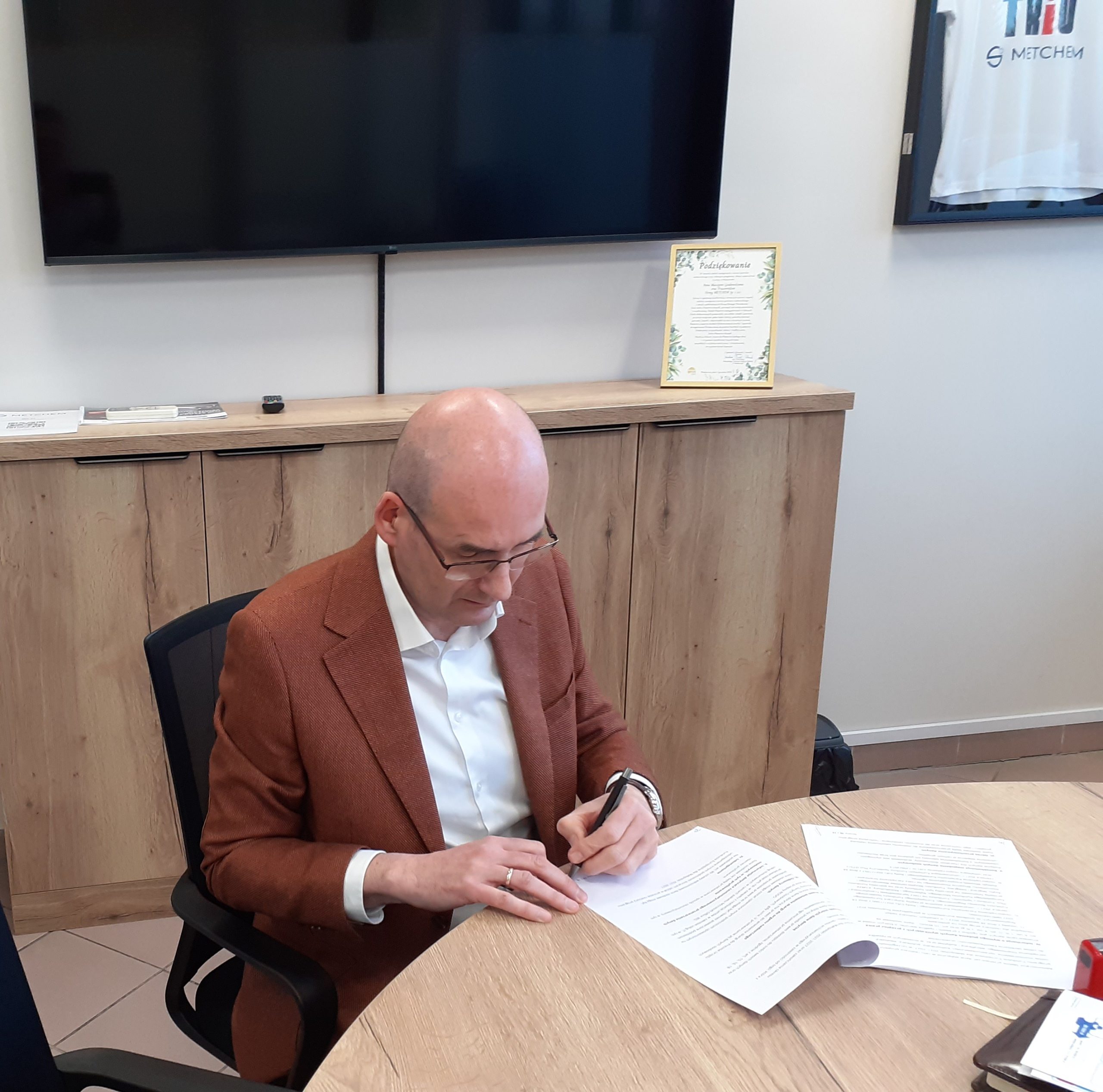 Mężczyzna w okularach i brązowej marynarce podpisuje dokumenty przy stole.