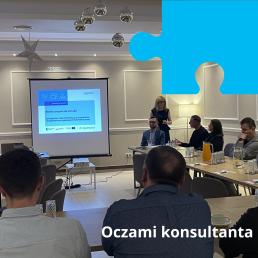 Grupa ludzi siedzi na sali konferencyjnej, patrząc na ekran z prezentacją pod tytułem: 