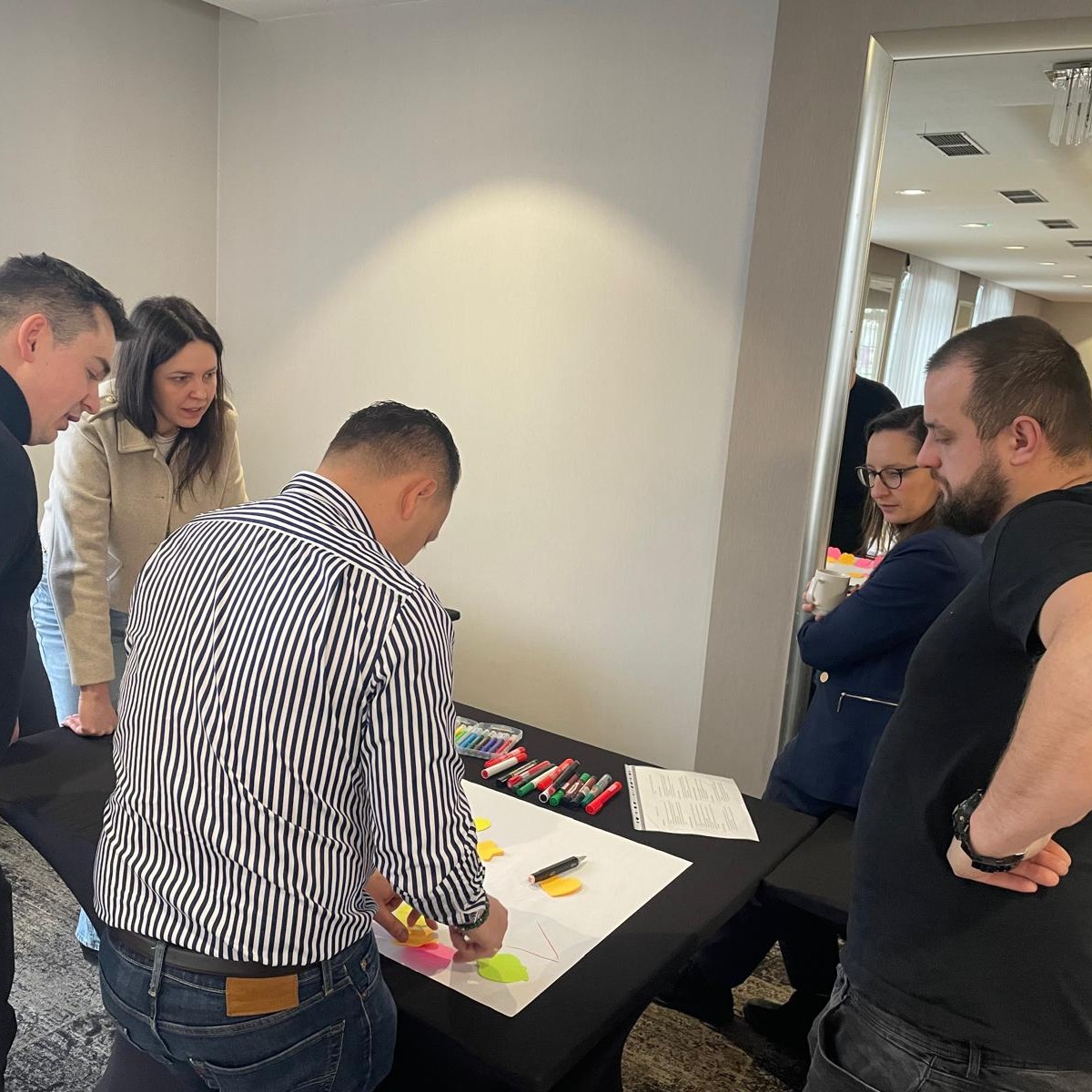 Trzech mężczyzn i dwie kobiety pochylają się nad stołem i przyklejają kolorowe karteczki do białej płachty papieru.