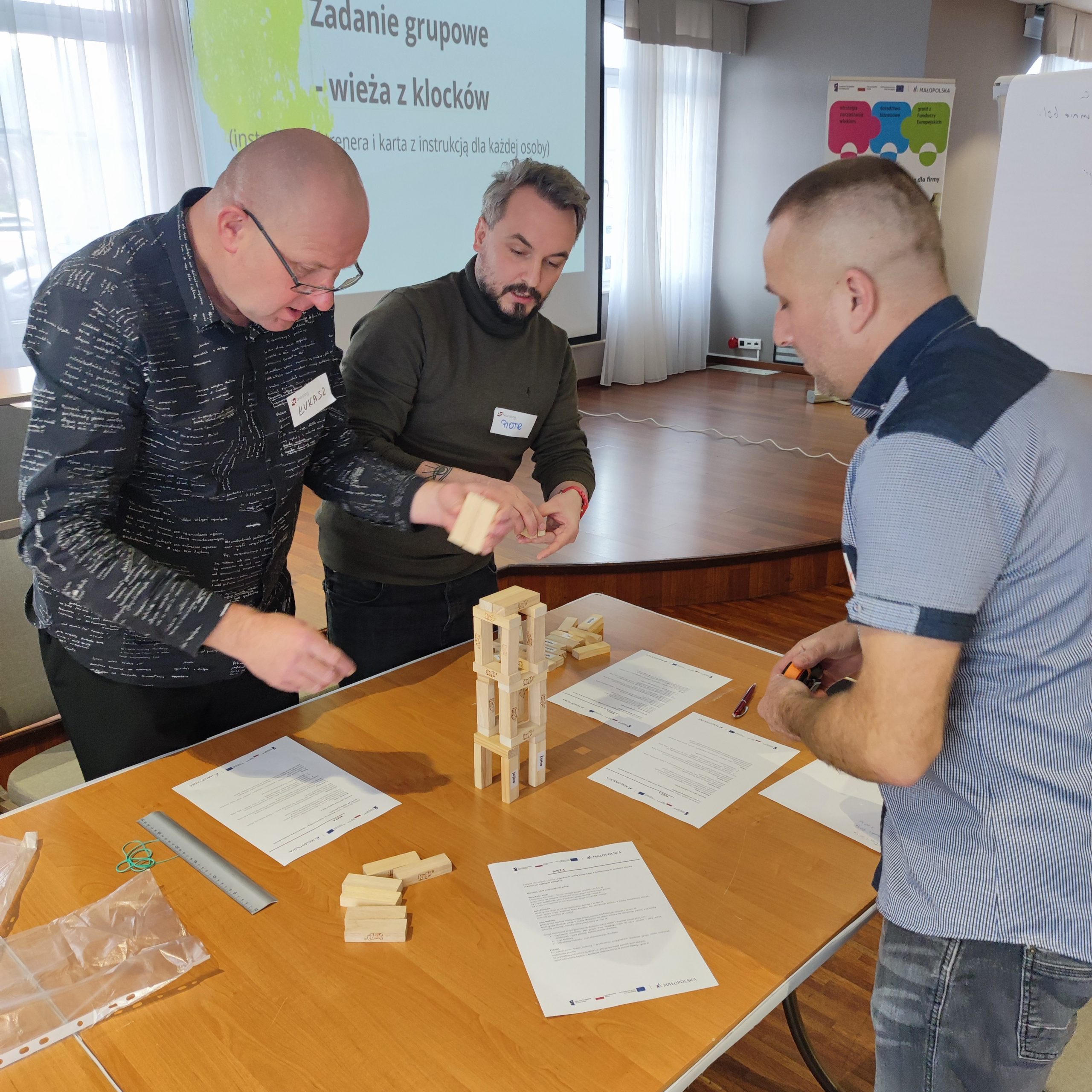 Trzech mężczyzn buduje wieżę z drewnianych klocków na szkoleniu.