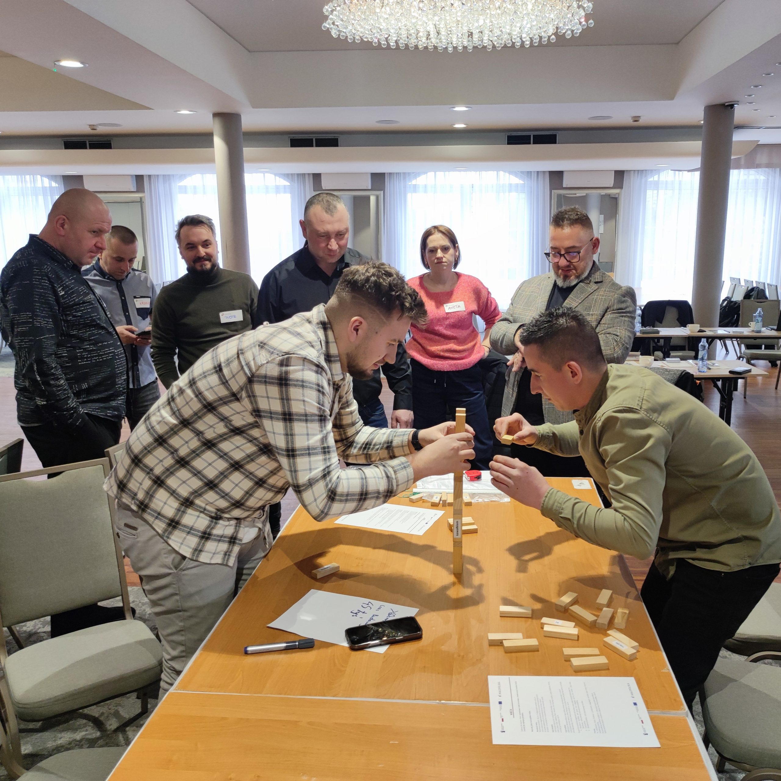 Dwóch mężczyzn buduje wieżę z klocków na szkoleniu, a grupa ludzi przygląda się im.