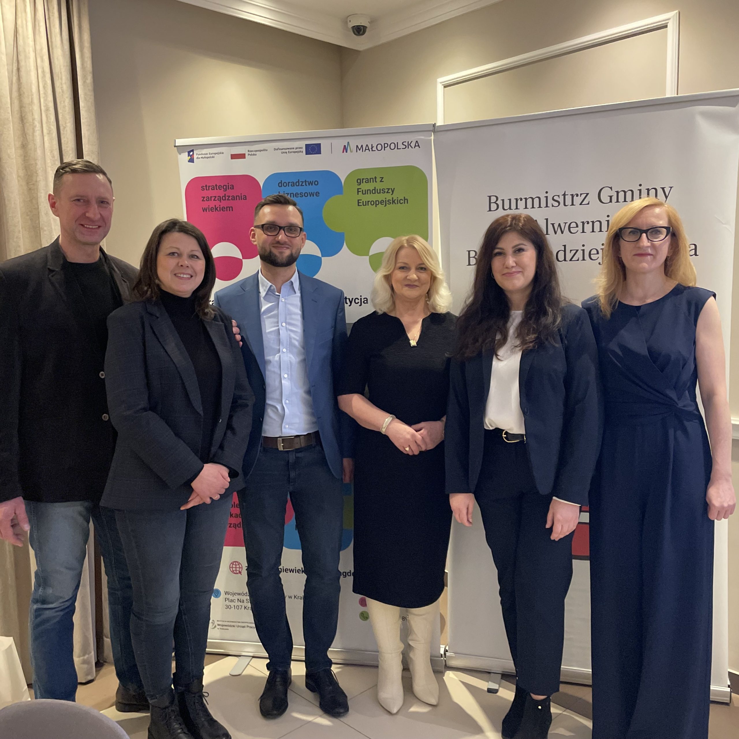 Cztery kobiety i dwóch mężczyzn w eleganckich ubraniach stoi na tle baneru projektu "Pracownicy - najlepsza inwestycja dla firmy" oraz baneru Burmistrza Gminy Alwernia