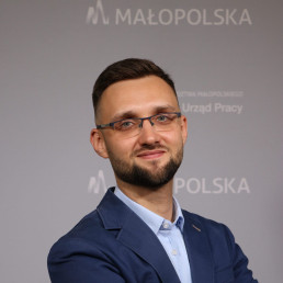 Młody mężczyzna w okularach, ciemnymi krótkimi włosami, z zarostem. Ma na sobie elegancką koszulę oraz marynarkę. W tle napisy Małopolska, Wojewódzki Urząd Pracy w Krakowie