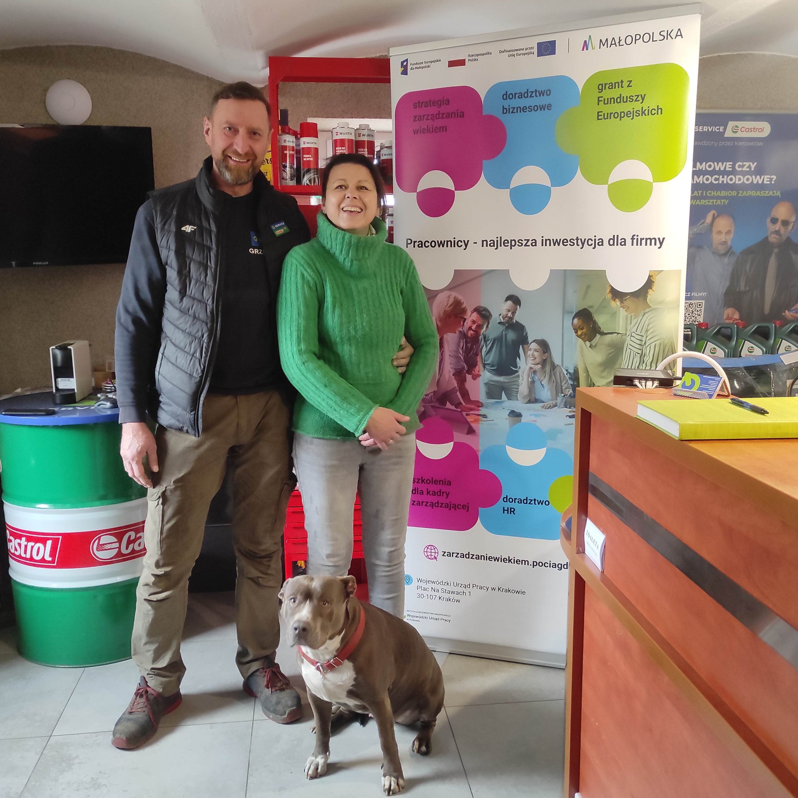 Kobieta i mężczyzna stoją uśmiechnięci na tle ścianki z napisem: "Pracownicy - najlepsza inwestycja dla firmy". Przed nimi siedzi pies.
