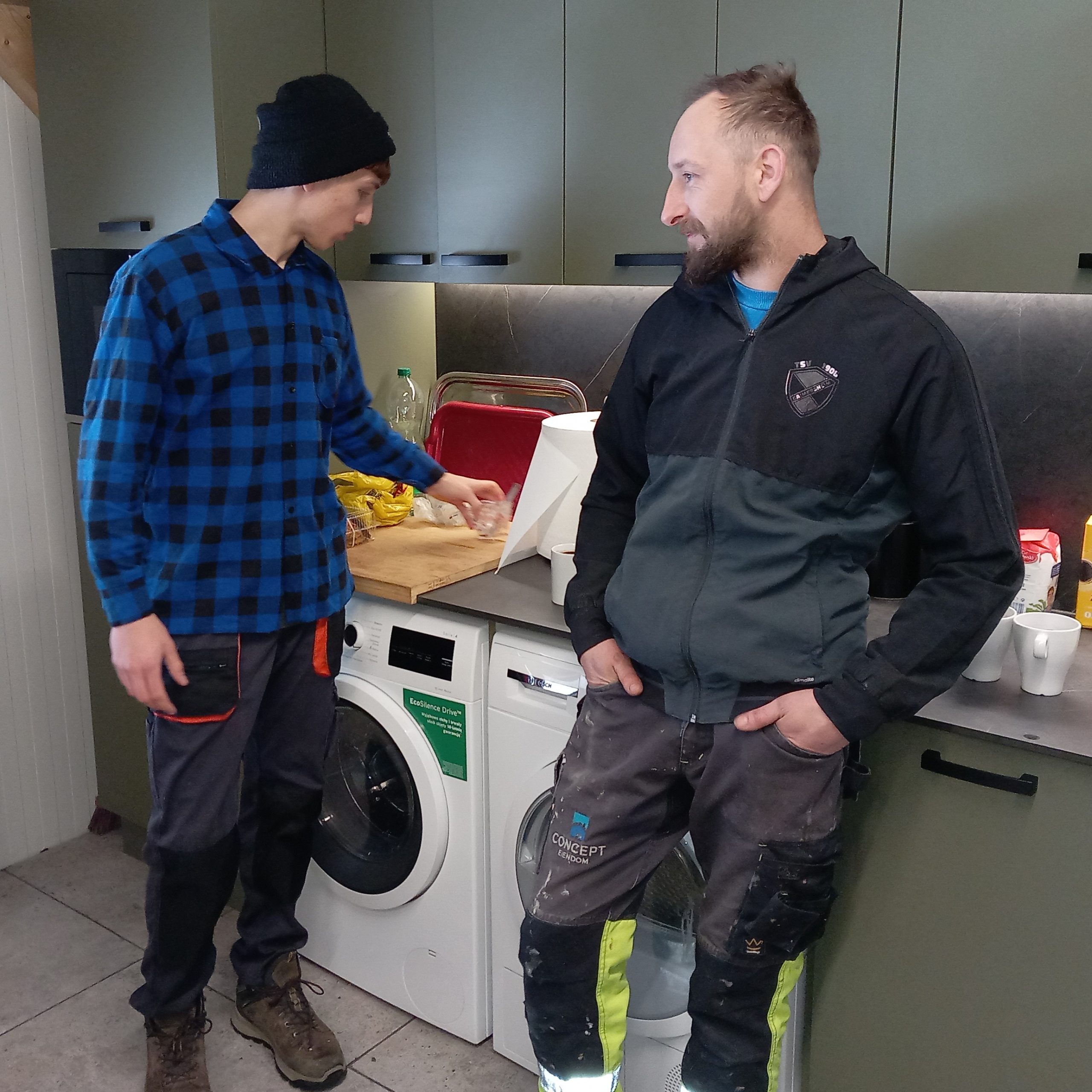 Dwóch młodych mężczyzn w roboczych uraniach stoi w kuchni. Jeden opiera się o blat, na którym leżą akcesoria kuchenne, drugi odstawia szklankę.