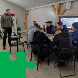 Grupa młodych mężczyzn siedzi przy stole. Rozmawiają ze stojącym naprzeciw nich mężczyzną w średnim wieku.