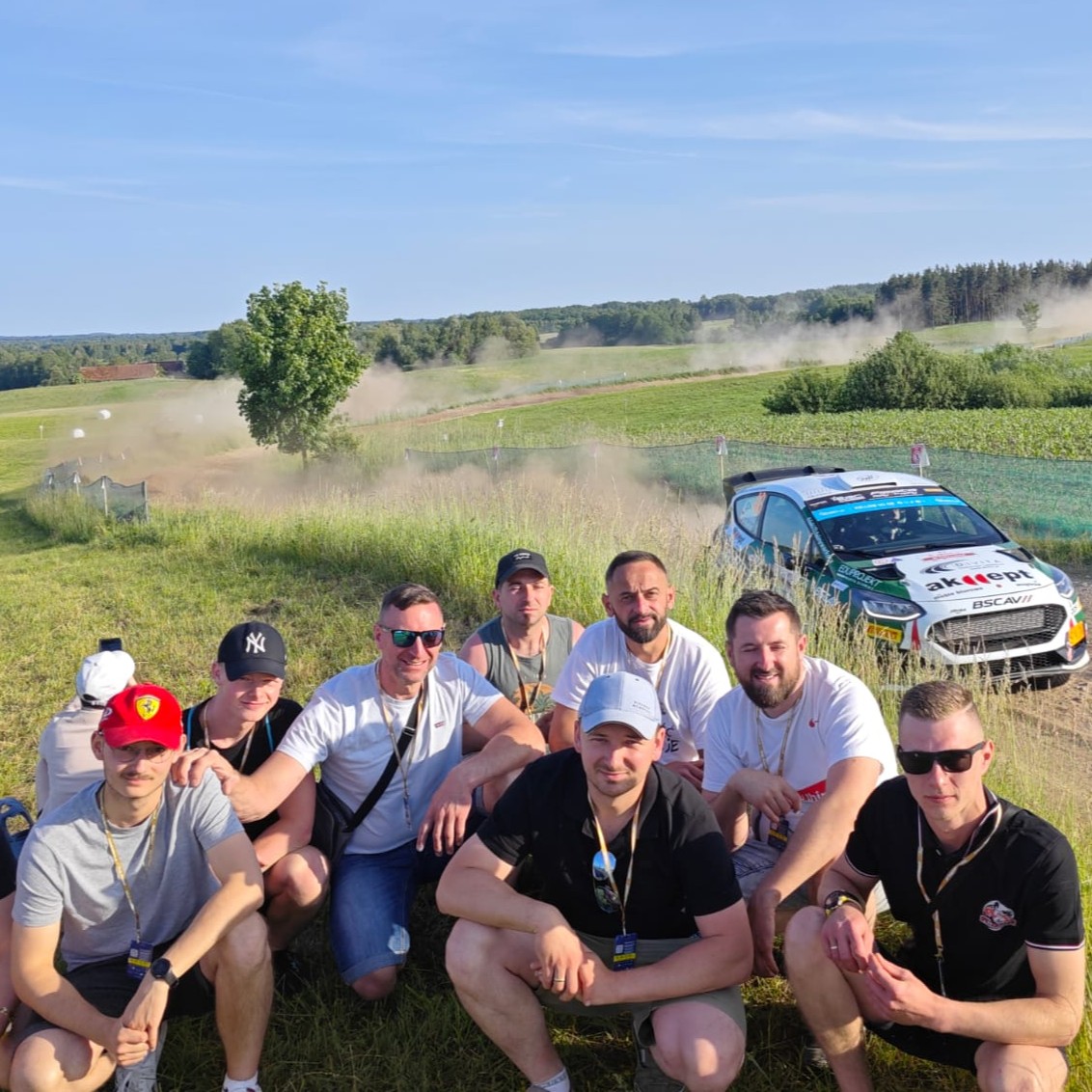 Grupa mężczyzn pozuje do zdjęcia w plenerze, na tle jadącego samochodu rajdowego.
