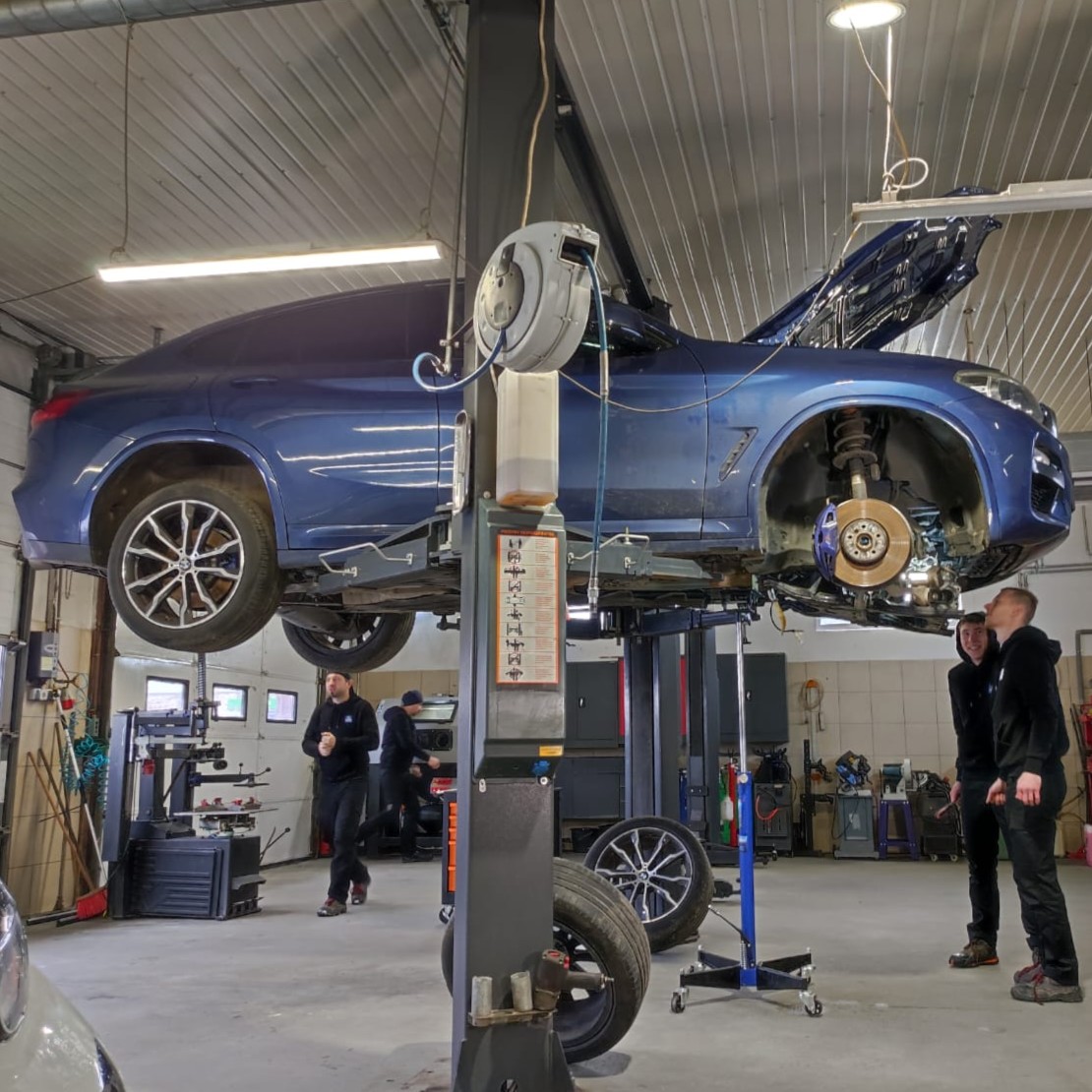 Dwóch młodych mężczyzn stoi przy niebieskim samochodzie na podnośniku. W tle warsztatu krzątają się dwaj inni mężczyźni.