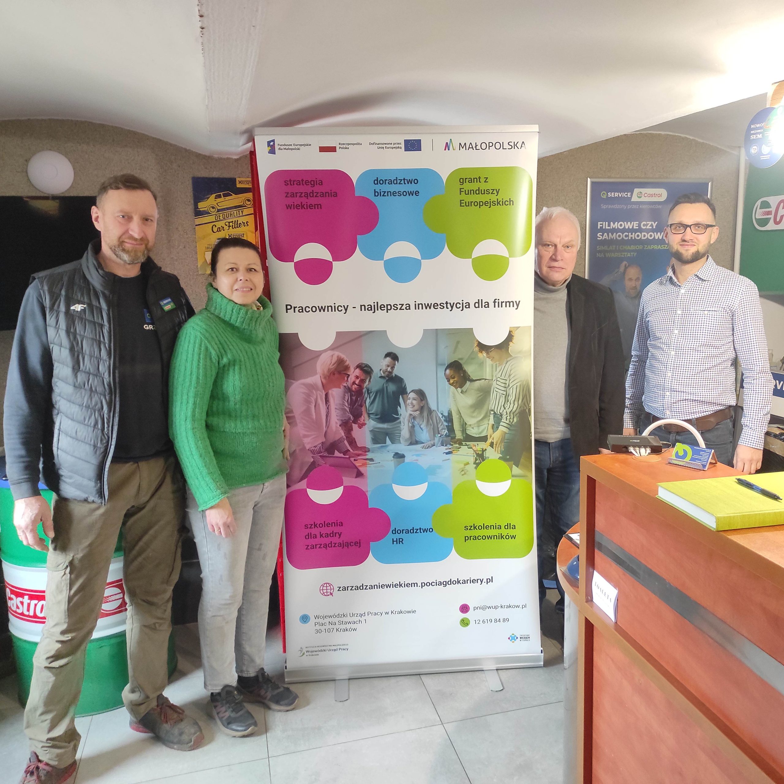 Trzech mężczyzn i kobieta stoją przy ściance z napisem: "Pracownicy - najlepsza inwestycja dla firmy". W tle wnętrze biura z akcesoriami samochodowymi.