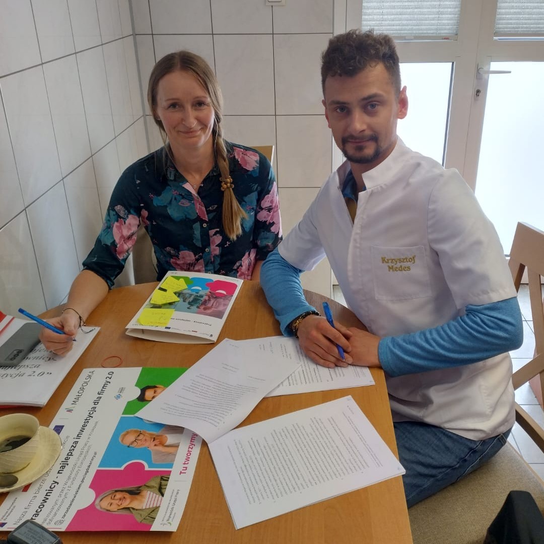 Uśmiechnięta kobieta z długim warkoczem i młody mężczyzna w białym kilku siedzą przy stole, na którym rozłożone są dokumenty.