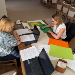 Dwie kobiety siedzące przy stole, pochylają się nad dokumentami, na stole leżą dwa laptopy