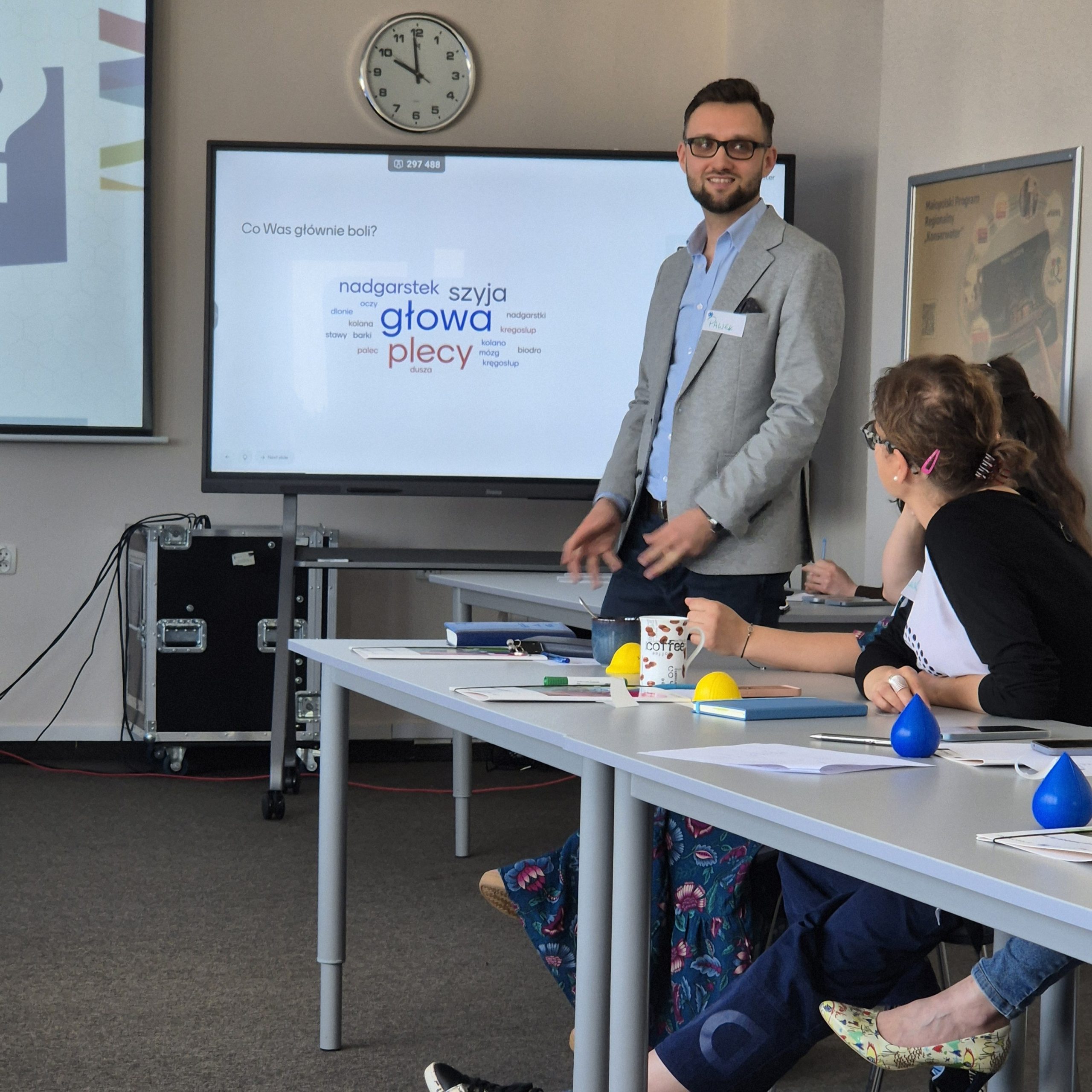 Młody mężczyzna stoi przy tablicy interaktywnej i mówi do ludzi siedzących przy stołach.