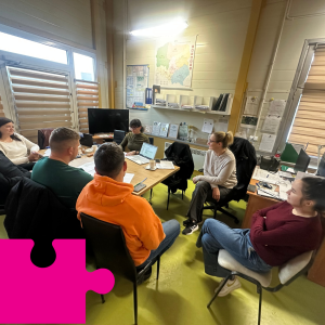 Grupa osób siedzących przy stole, rozmawia, na stole laptop oraz dokumenty